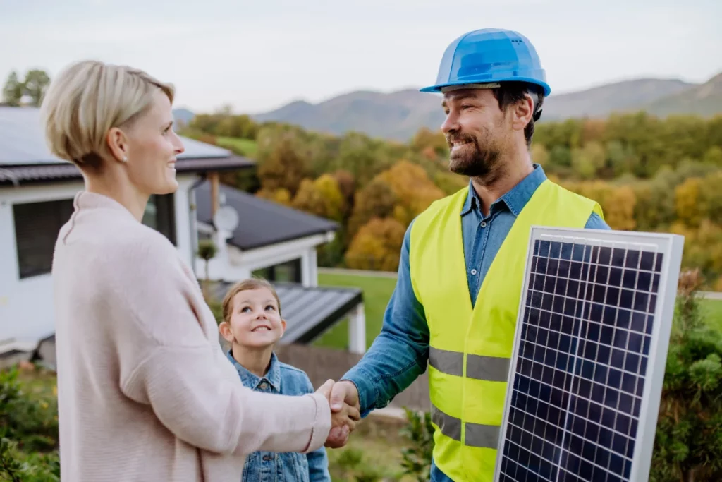 Arbeiter mit Warnweste und PV Modul in der Hand schüttelt Mutter mit Kind die Hand